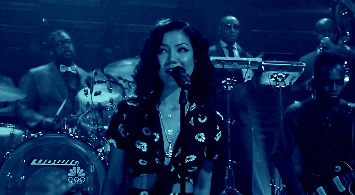 A blueish filter picture of Jhene Aiko in a black dress singing The Worst.